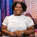 A smiling woman wearing a white shirt sits behind a desk with her hands resting on the desk in front of her.