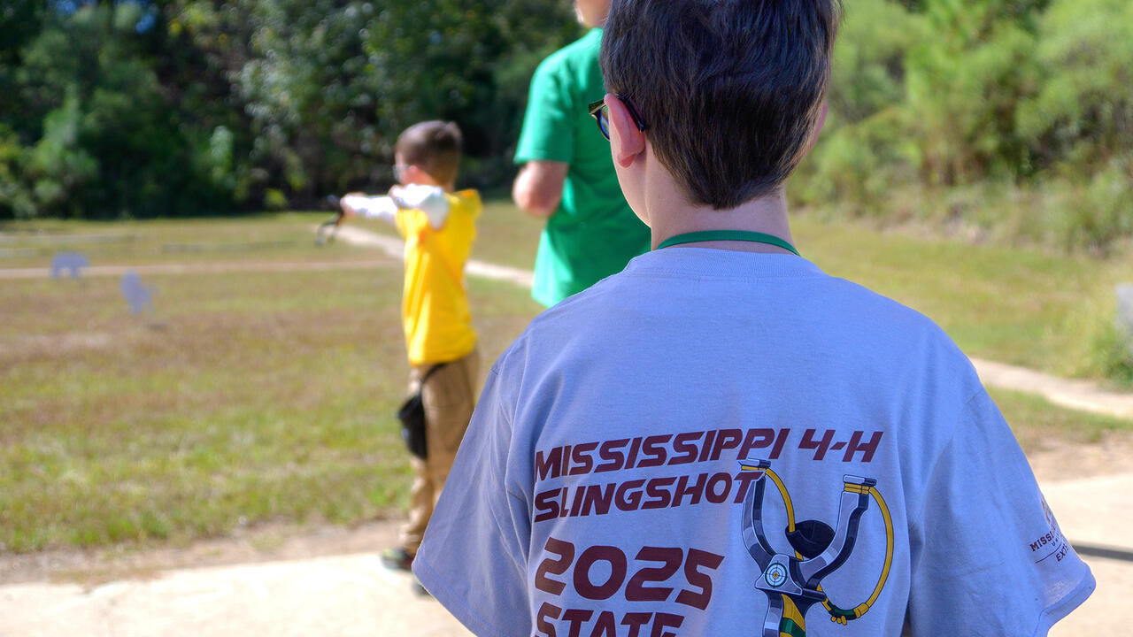 In the foreground, a boy pictured from behind wearing a T-shirt reading “Mississippi 4-H Slingshot 2025 State Championship” looks on as another boy aims at a target with a slingshot.