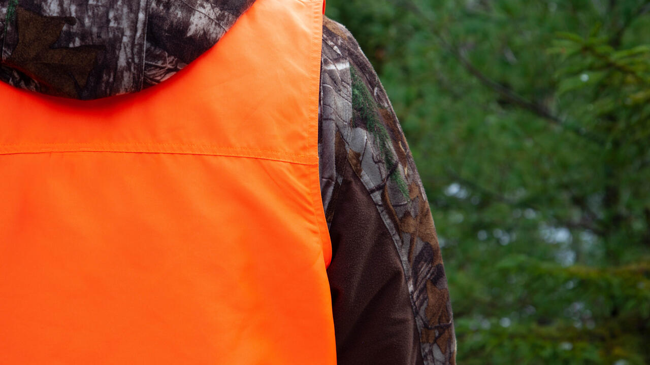 The back of a hunter wearing a camouflage jacket and an orange hunting vest