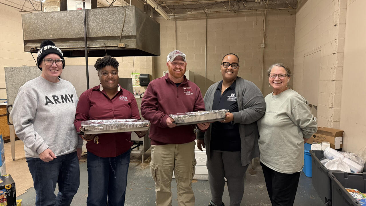 A group of five people standing together in an industrial-style room, with two of them holding large foil-covered food pans. Boxes, supplies, and equipment are visible around the room.