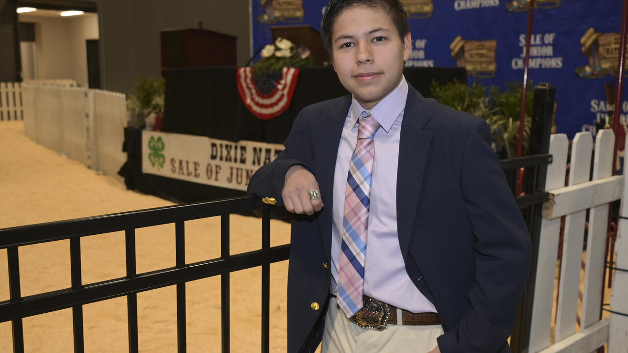 A person in a blazer and tie leans on a fence inside the Dixie National Sale of Junior Champions arena.