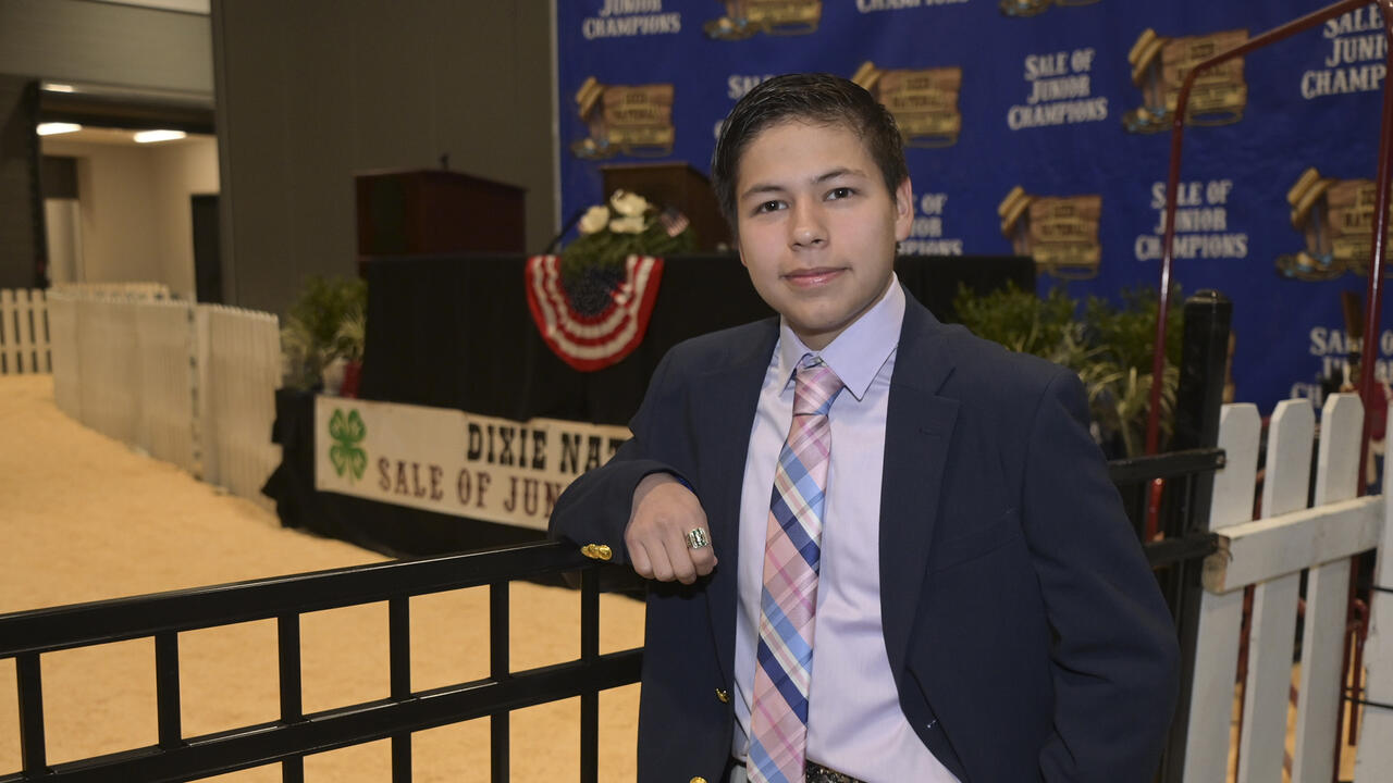 A person in a blazer and tie leans on a fence inside the Dixie National Sale of Junior Champions arena.