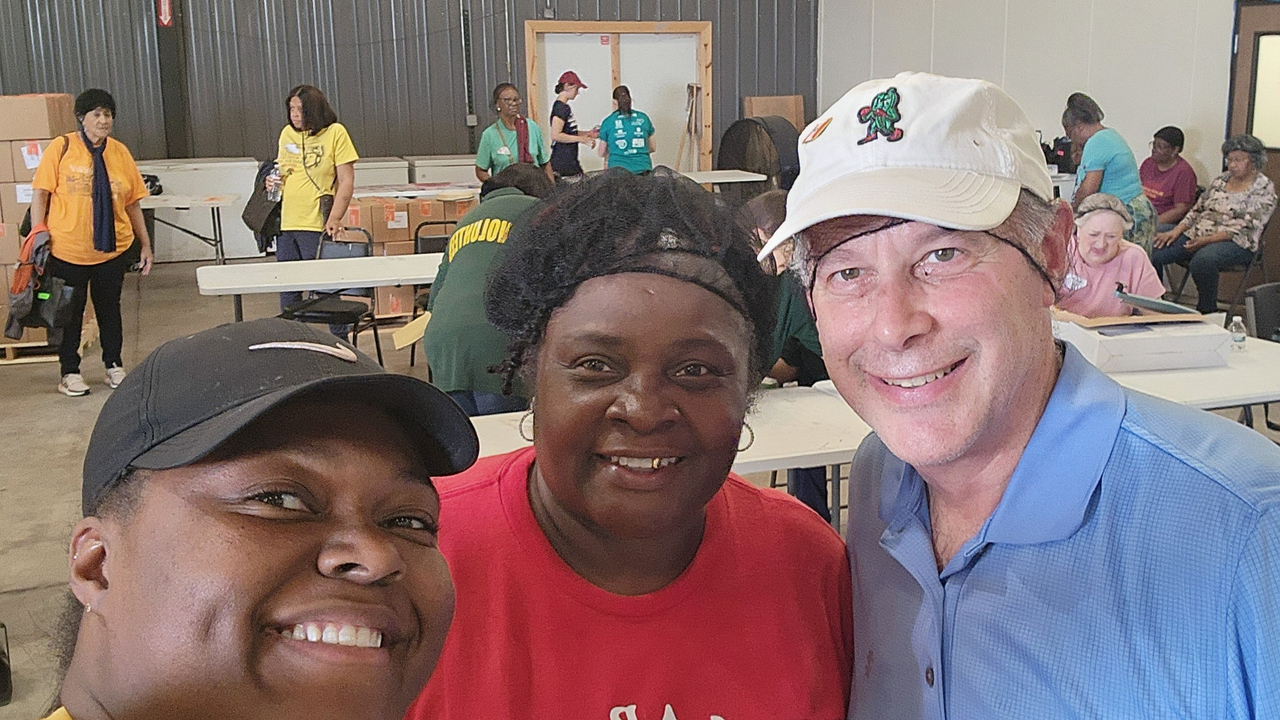 Three people pose for the camera in a church auditorium with others in the background as they prepare to distribute food.
