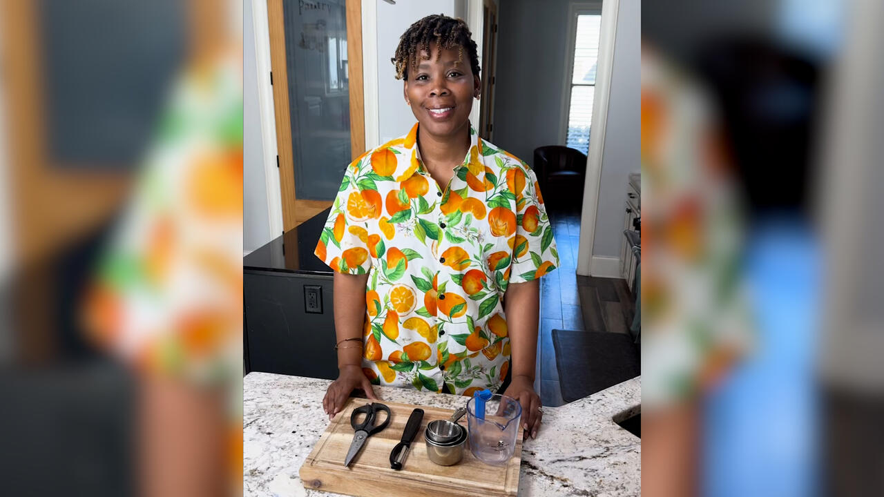 A woman stands in a kitchen with a cutting board, kitchen scissors, a vegetable peeler, and measuring cups on the counter in front of her.