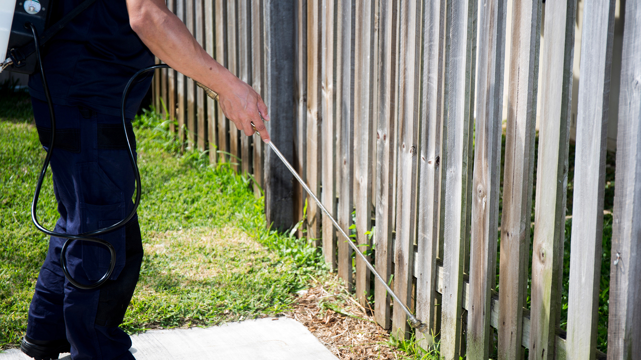 A person uses a wand to spray a fence outdoors.
