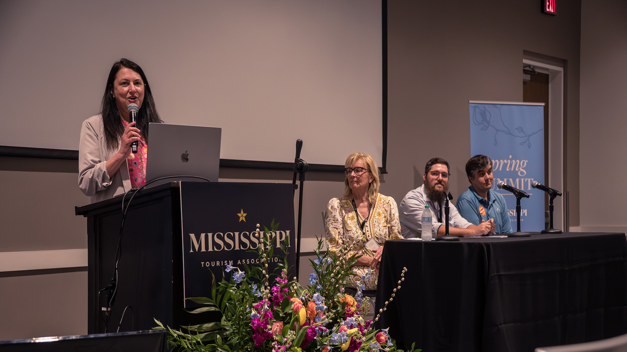 A speaker at a Mississippi Tourism Association podium addresses a panel seated behind a table in an auditorium.