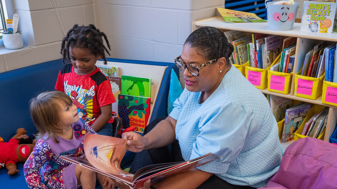 An adult reads from a picture book to two young children seated in a classroom reading corner.