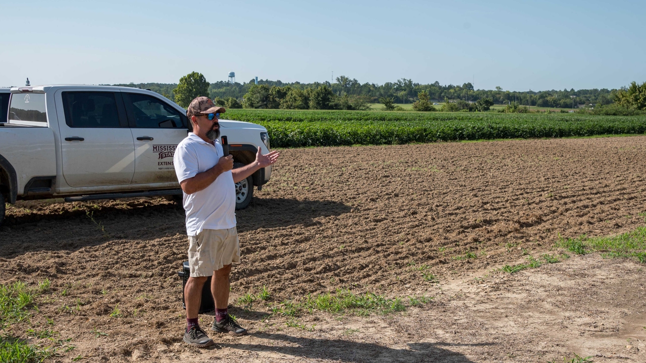A speaker stands in front of research plots with a Mississippi State University Extension pickup truck in the background.
