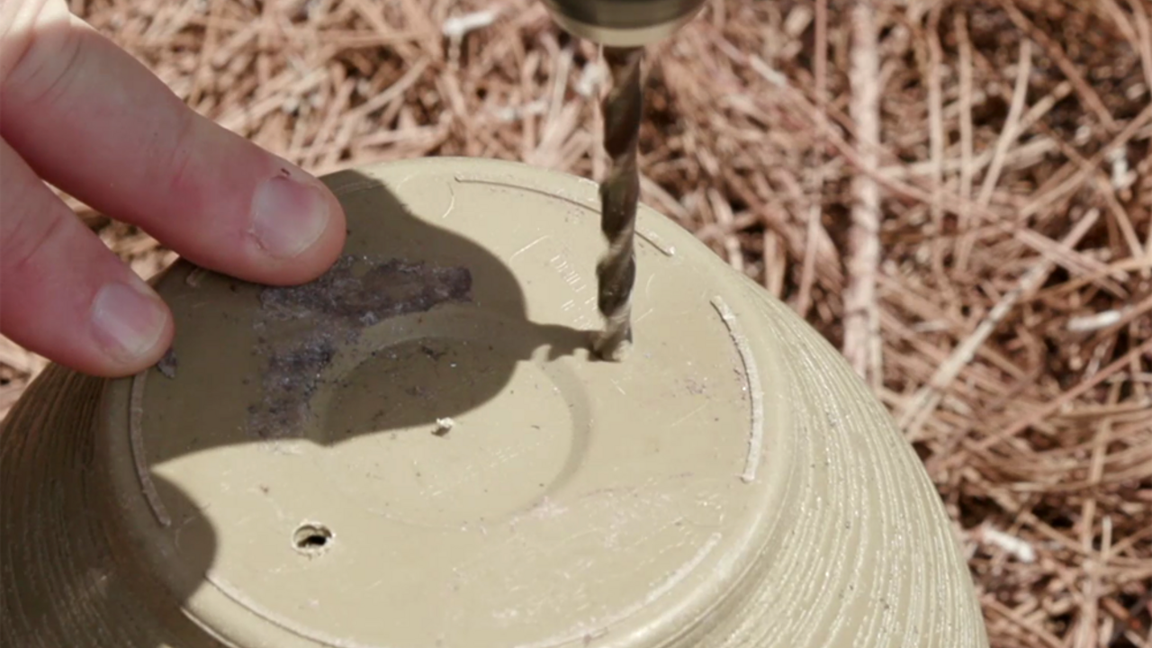 Fingers steady an upside-down flower pot as a drill bit pierces the bottom to make drainage holes. 