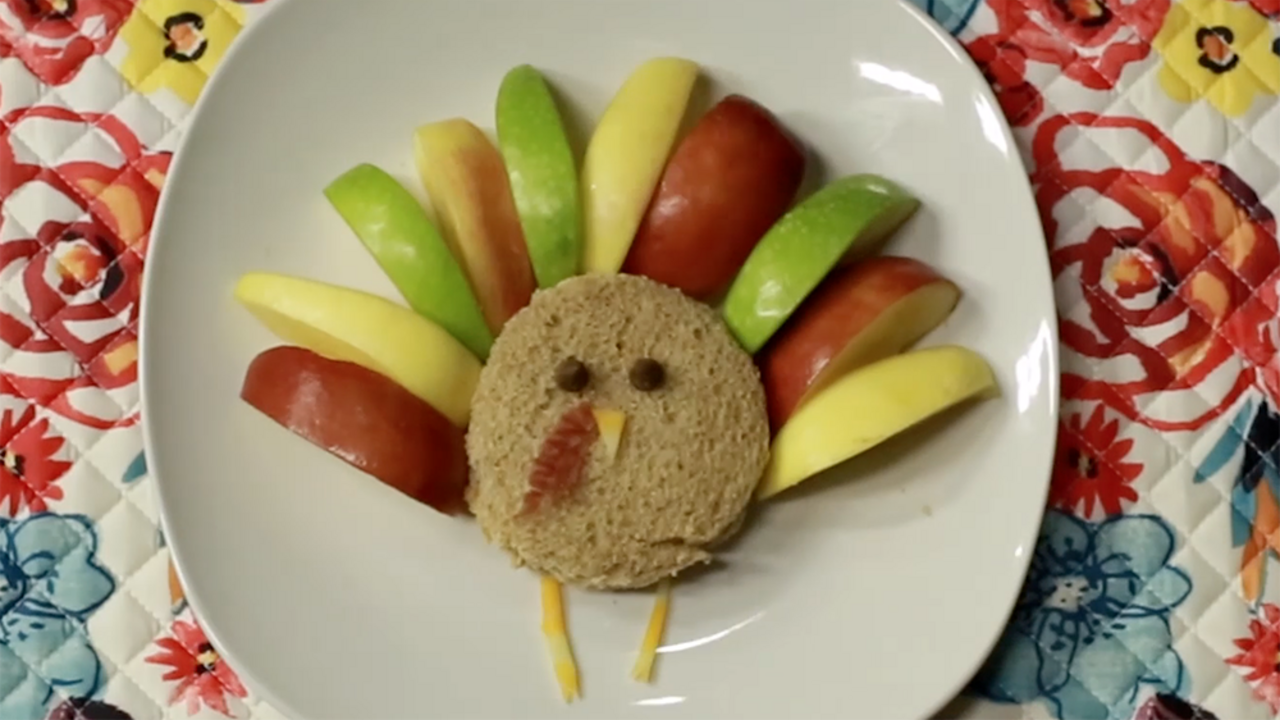 A round peanut butter and jelly sandwich sits on a white plate. It is decorated to look like a turkey, with green, red, and yellow apple slices for feathers, chocolate chips for eyes, cheese for a nose and legs, and a small bit of bacon for a wattle. The plate sits on a multicolored floral placemat. 