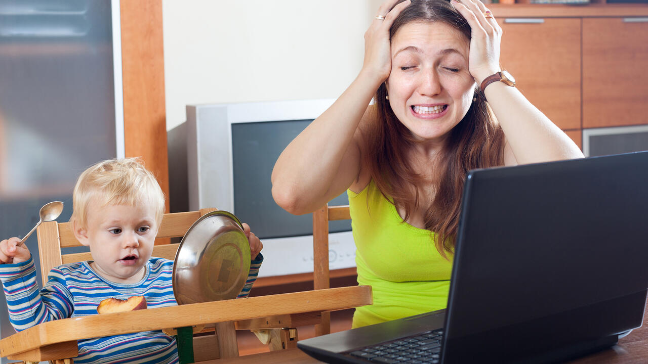 A young mother grabs her head in frustration while working from home on a laptop computer as her baby looks on. 