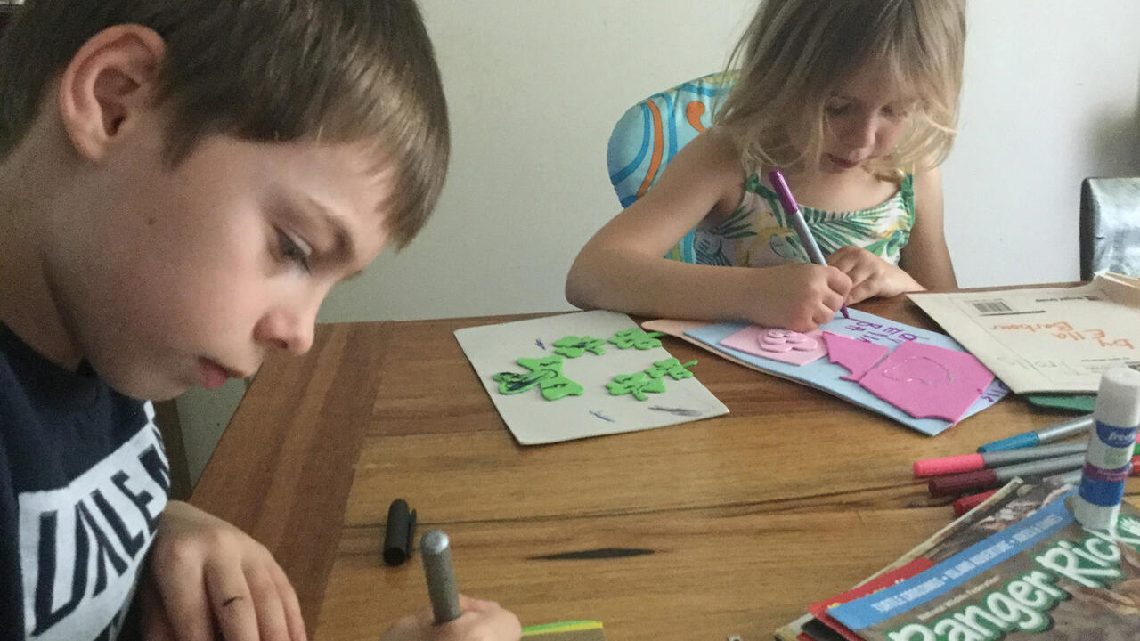 A boy and girl sit at a wooden table and use markers to draw on different colored sheets of paper.