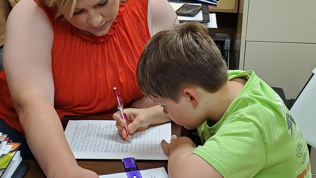 A woman looks on while a young boy writes on a piece of paper.