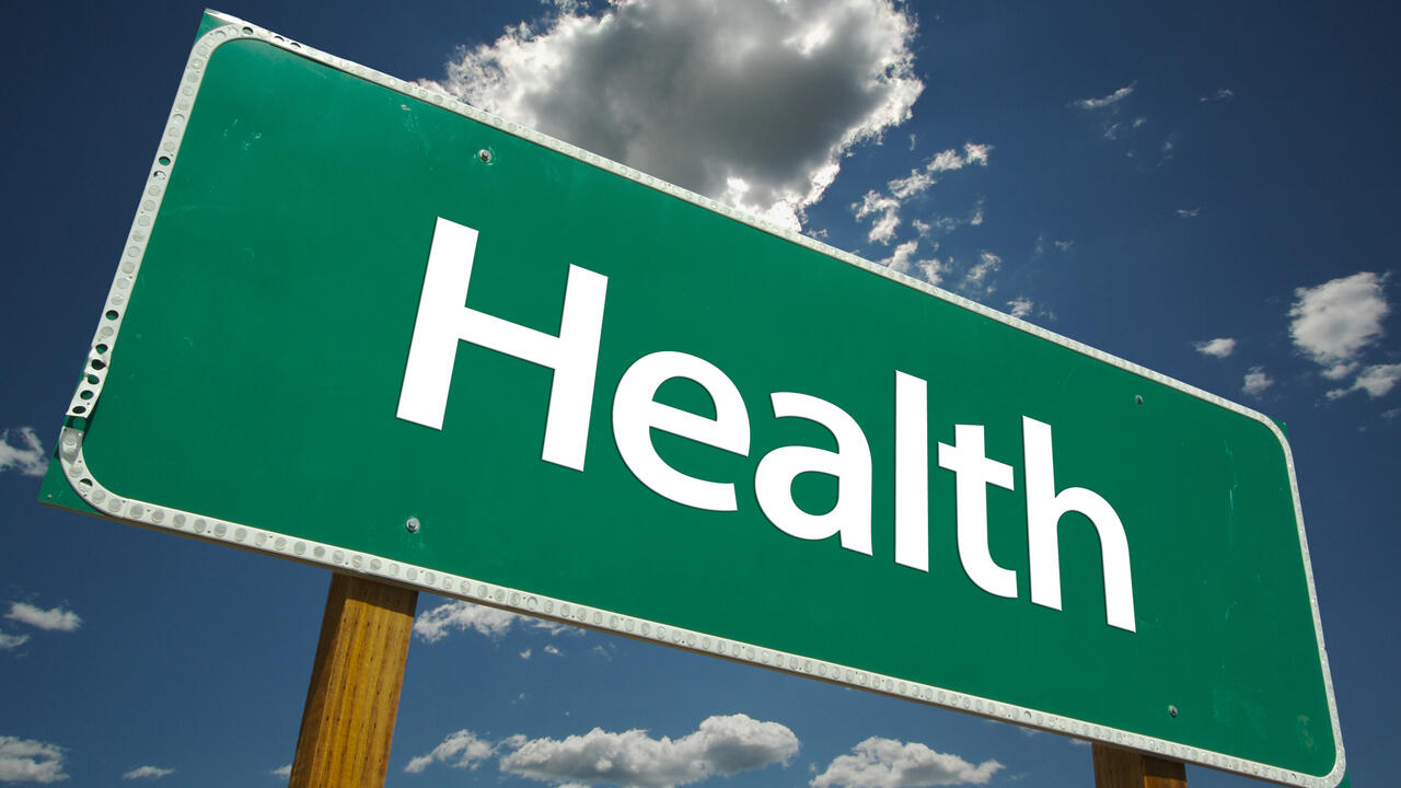 A green road sign with the word “Health” is pictured against a blue sky with white clouds.