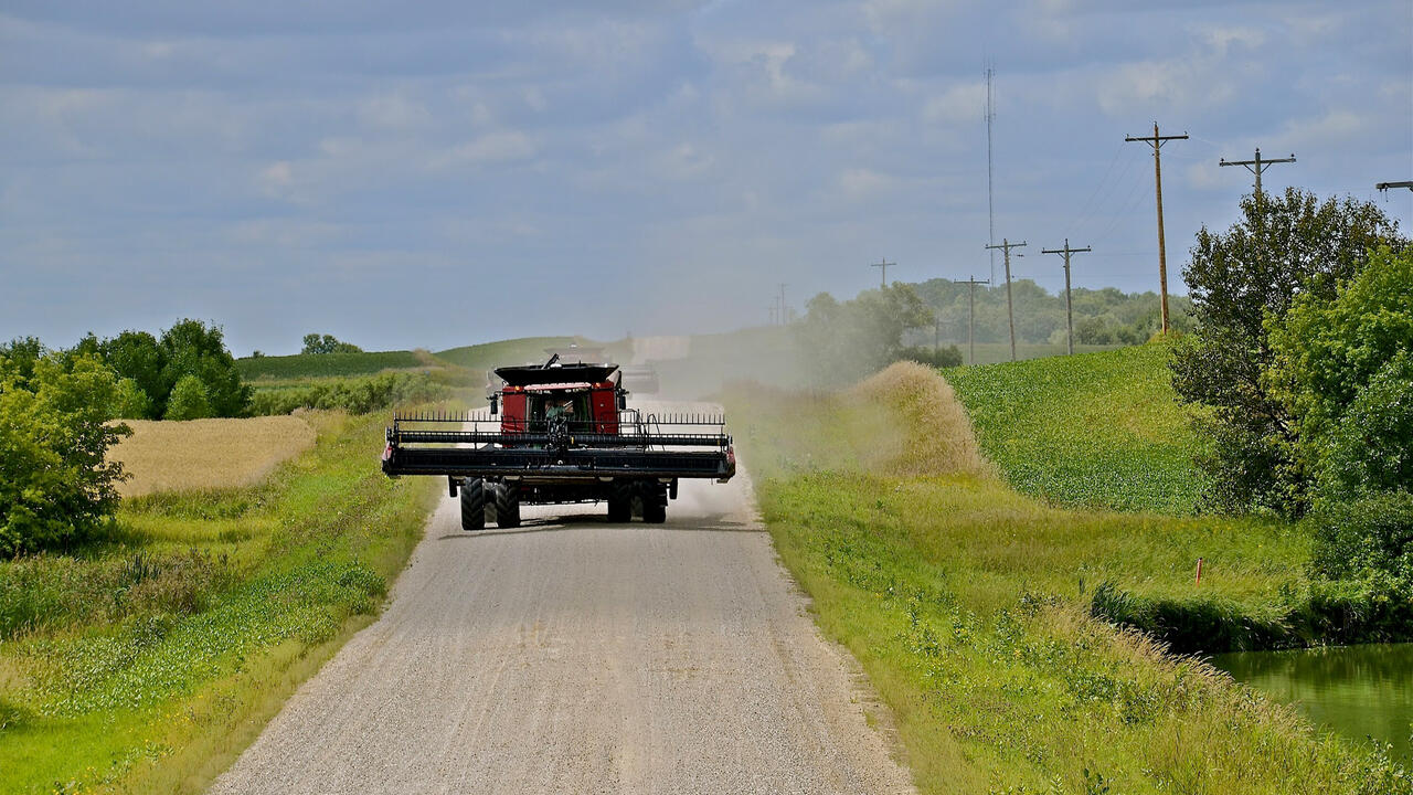 A red combine drives down a gravel road with farmland on both sides.