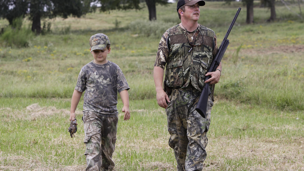 A man holding a shotgun and a boy dressed in camouflage walk in a grassy meadow.