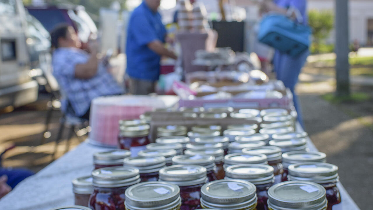 Rows of canning jars line a table at an open-air marketplace.