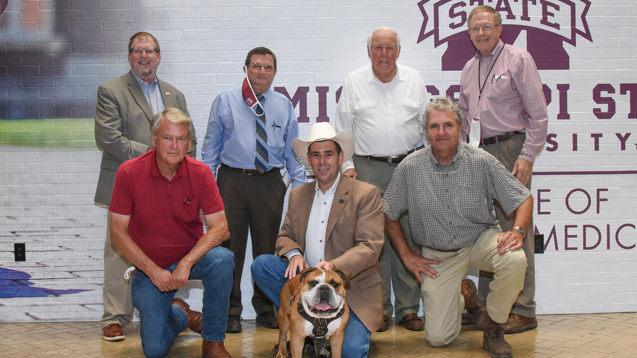 Seven men stand or kneel indoors in two rows, holding a bulldog in the center of the group.