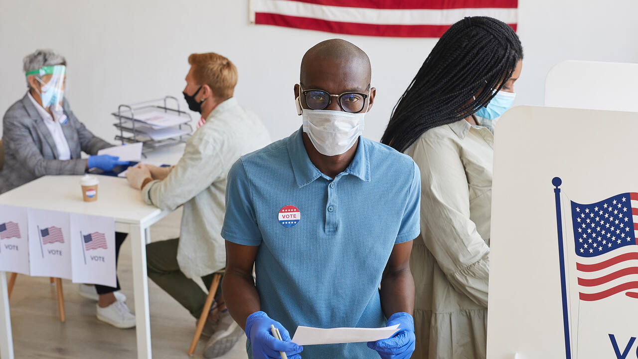 Voter wearing mask, standing by booth and looking at camera.