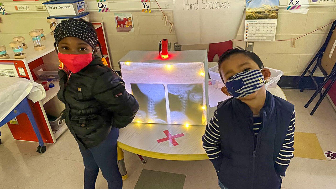 Two children in masks stand in front of an activity center in a classroom.