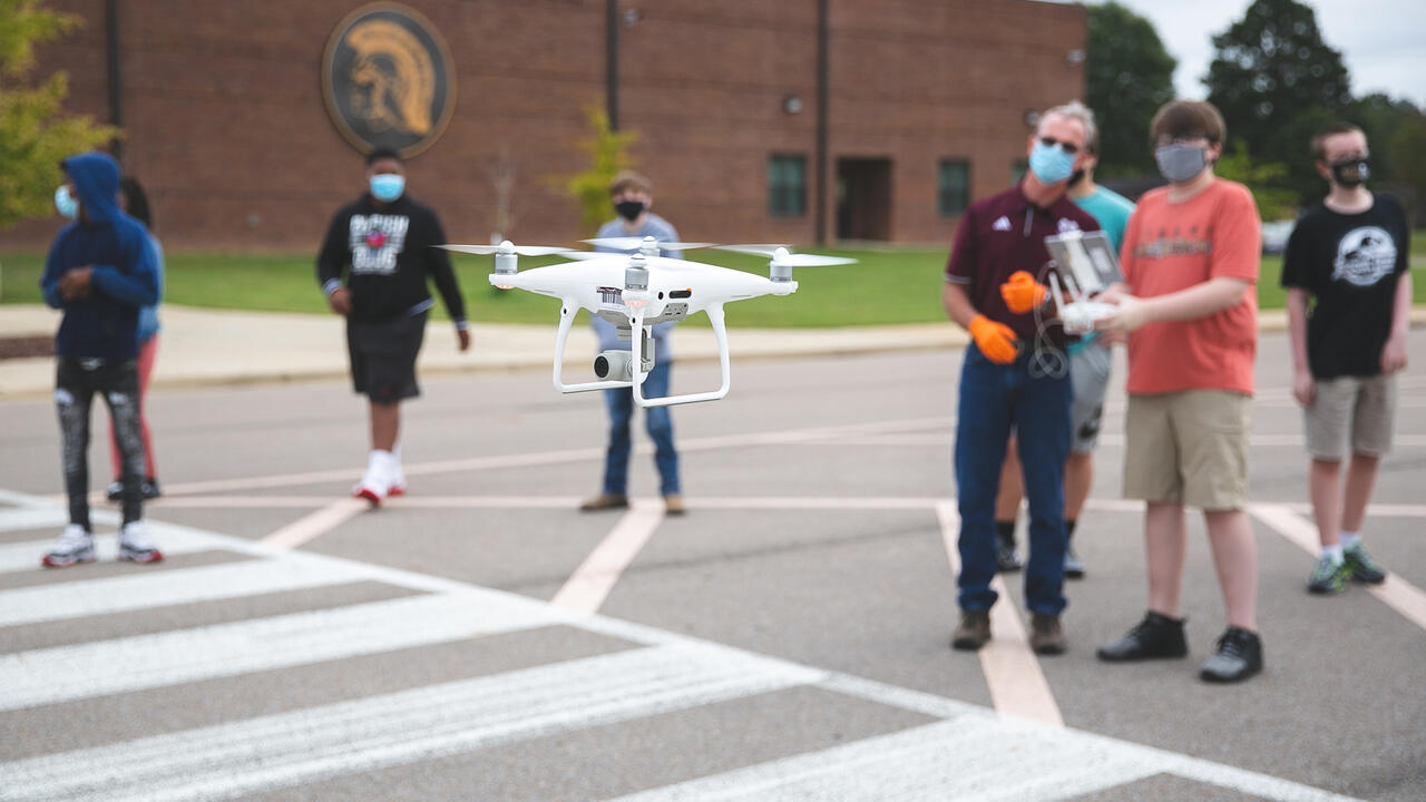 A drone in the foreground being controlled by young students in the background.