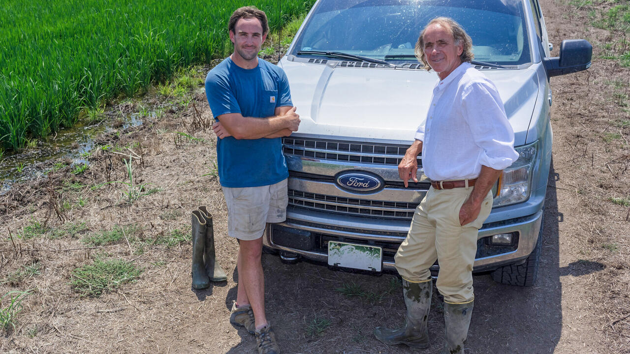 Two men standing in front of a pickup truck parked in a rice field.