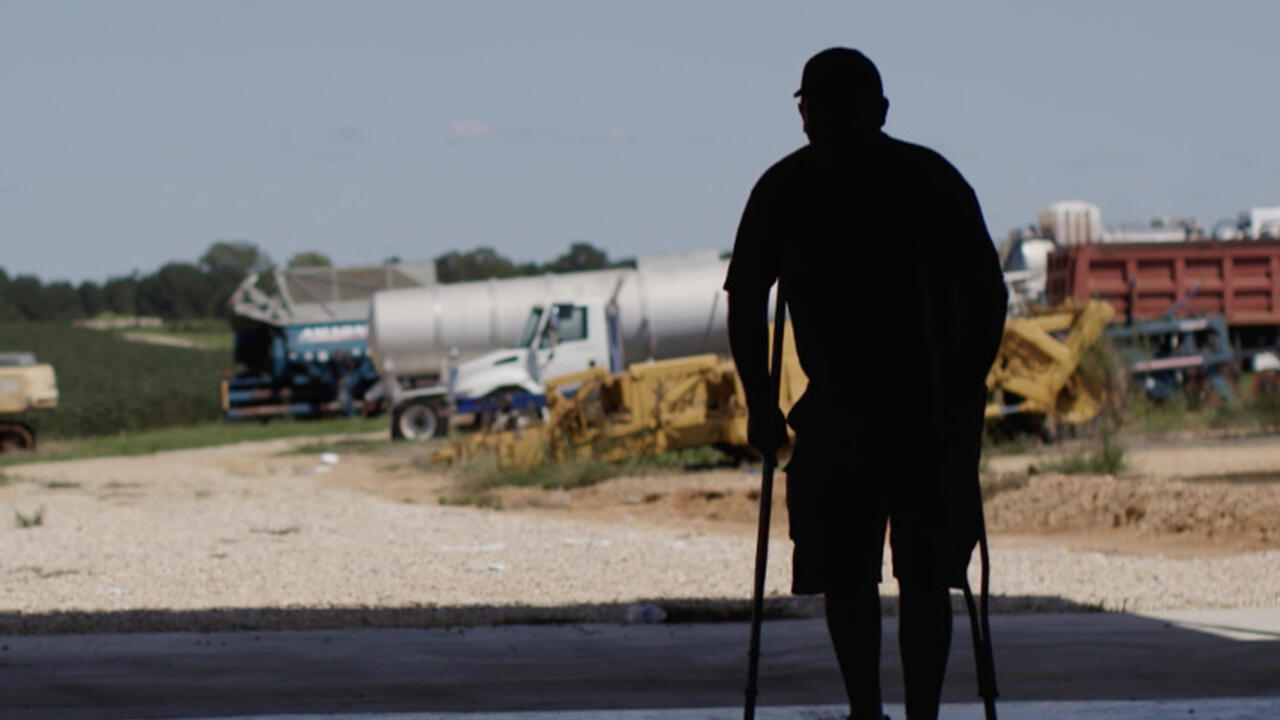 A man stands on crutches in silhouette against a background of farm equipment.