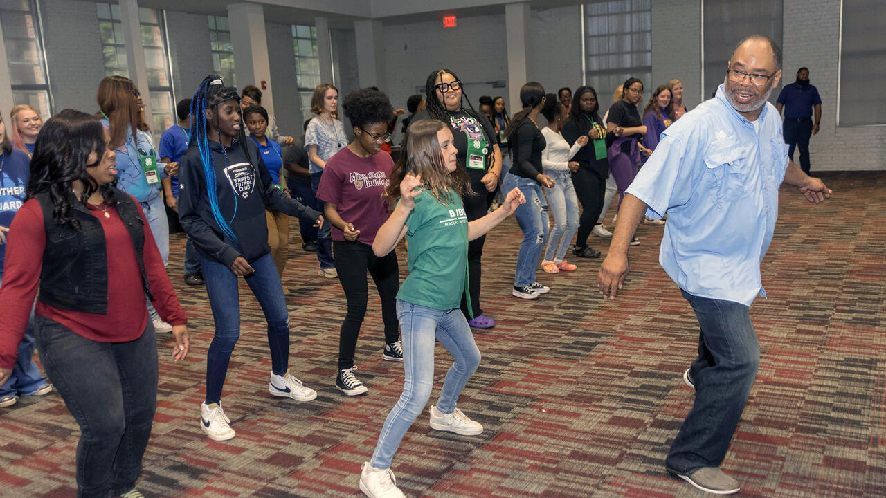 Man leads room full of teenagers in dance exercise.