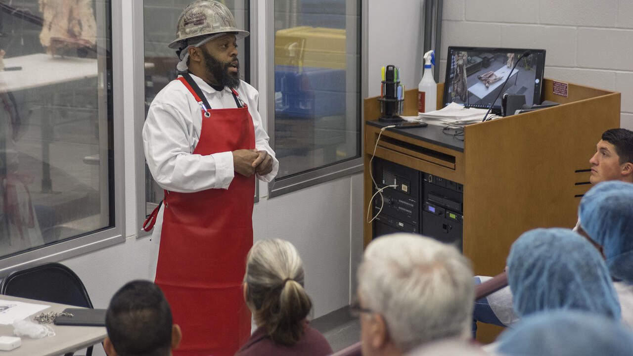 A man in a red apron speaks to a class in a lab.