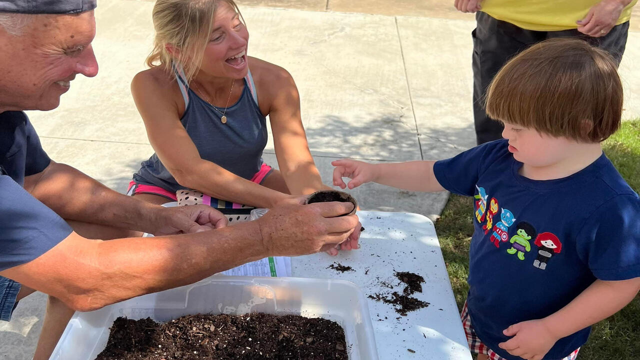 A man and a woman help a child plant seeds in a small pot of soil.
