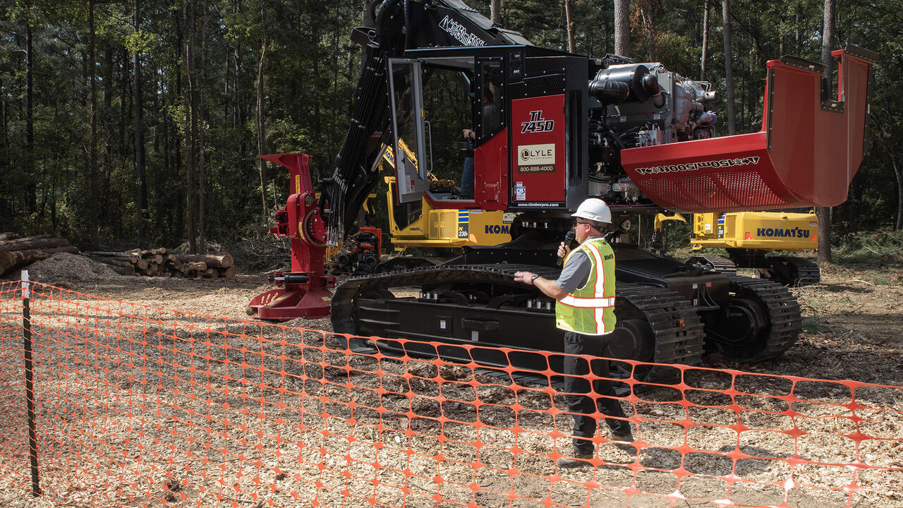 A man wearing a hard hat talking into a microphone in front of a piece of heavy equipment.
