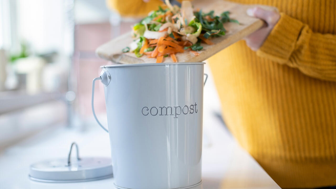 A woman scrapes vegetable scraps off a cutting board into a countertop compost bin.