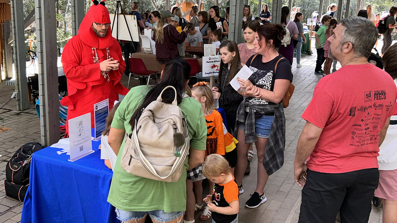 Several adults and children gather around an educational booth hosted by a man in a crawfish suit.