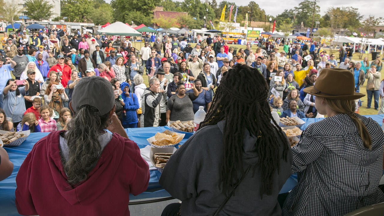 A large crowd gathers at a festival while a panel of judges tastes food.