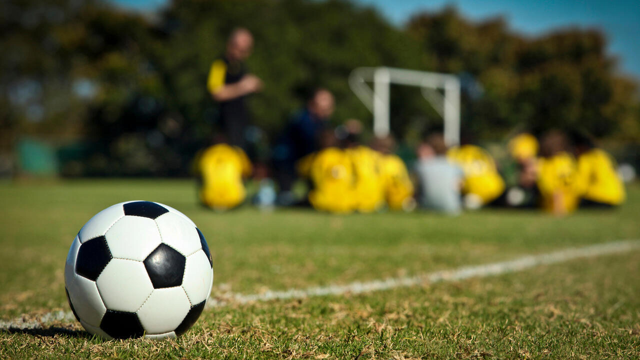 A soccer ball sits in the foreground with a soccer team and coach in the background.