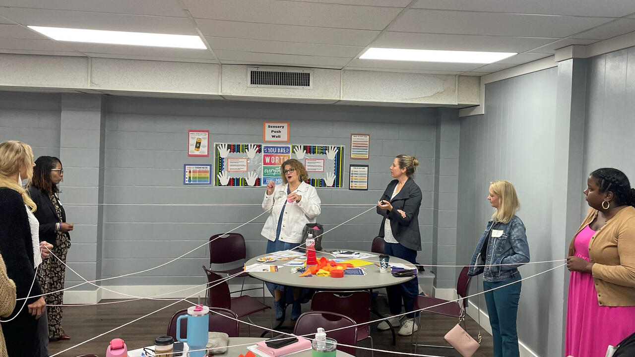 A group in a circle holds a white string that crisscrosses a classroom.
