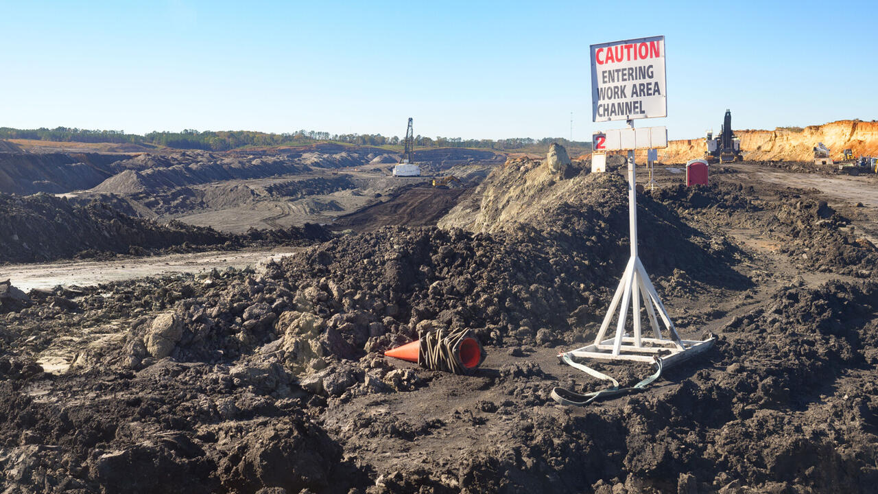 Wide shot of a surface coal mine featuring a layered landscape of exposed rock and soil, heavy machinery, and a sign that reads “caution, entering work area channel.