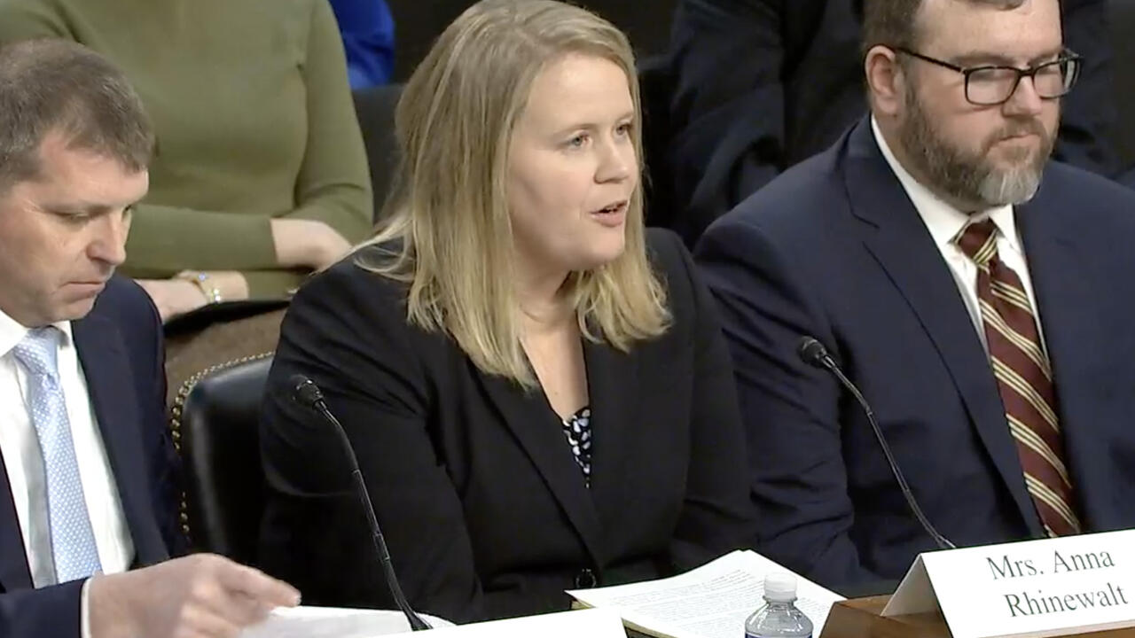 Woman seated and speaking into a microphone with men sitting beside her.