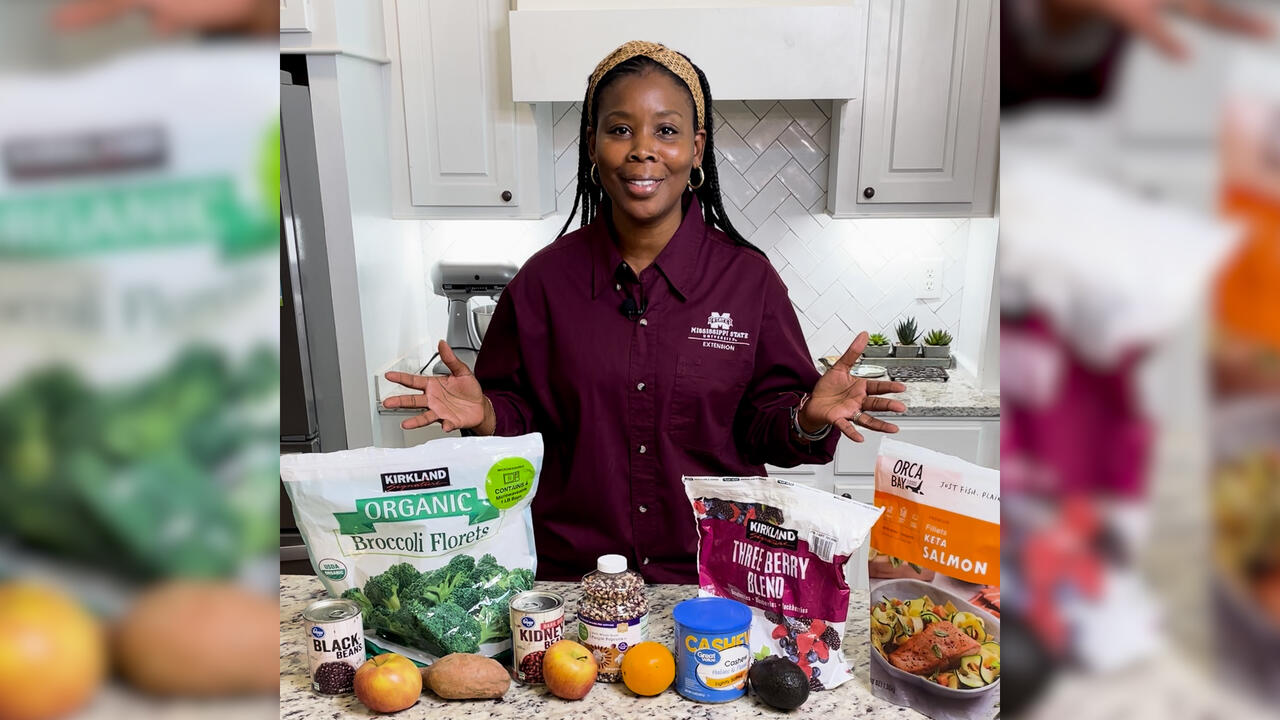 A woman stans in a kitchen with fruits, vegetables, fish, nuts and other foods on display.