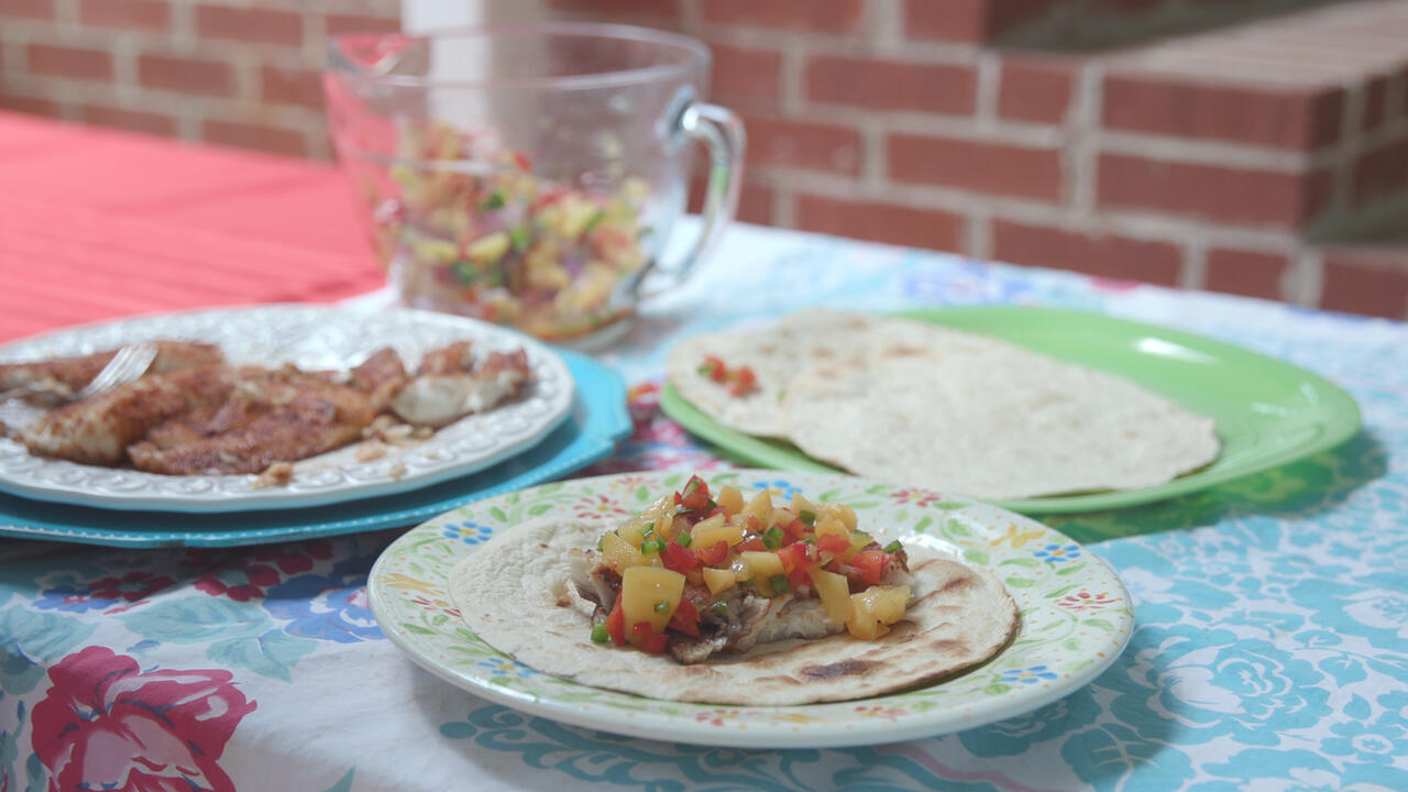 Three plates with tortillas, fish, and a completed grilled fish taco with peach salsa on a picnic table. 
