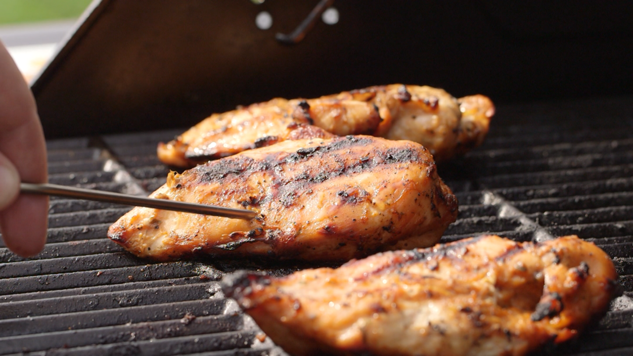 Hand with meat thermometer chicken middle of 3 chicken breasts on outdoor grill.