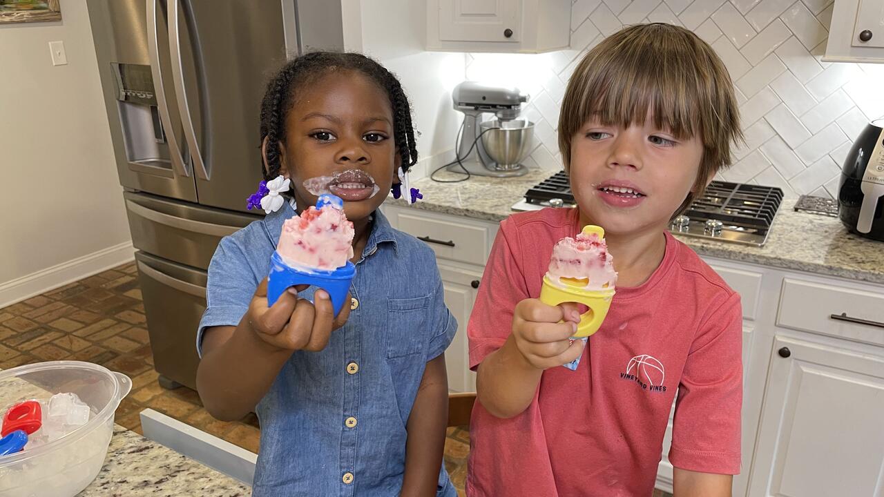 A girl and boy stand in a kitchen and hold up the popsicles they are eating.