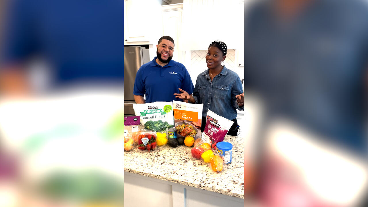 A man and woman stand behind a kitchen counter filled with fruits and vegetables.