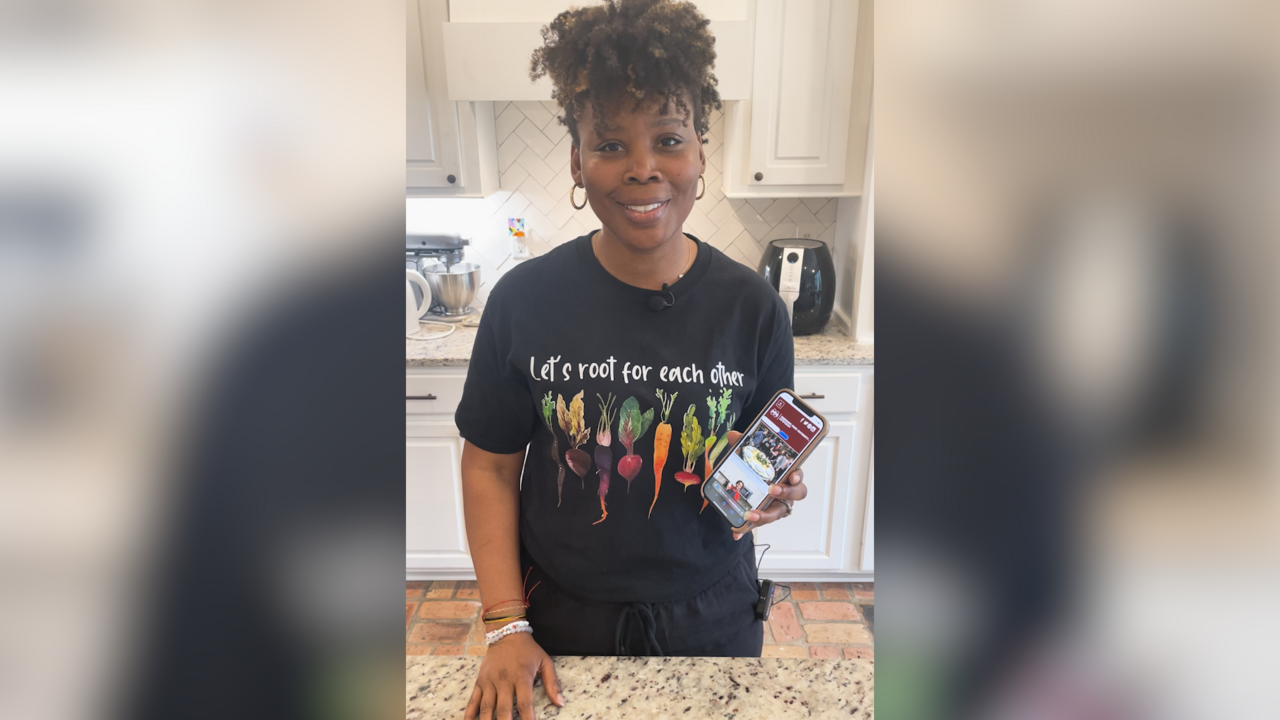 A woman stands in kitchen with a cell phone displaying a web page.