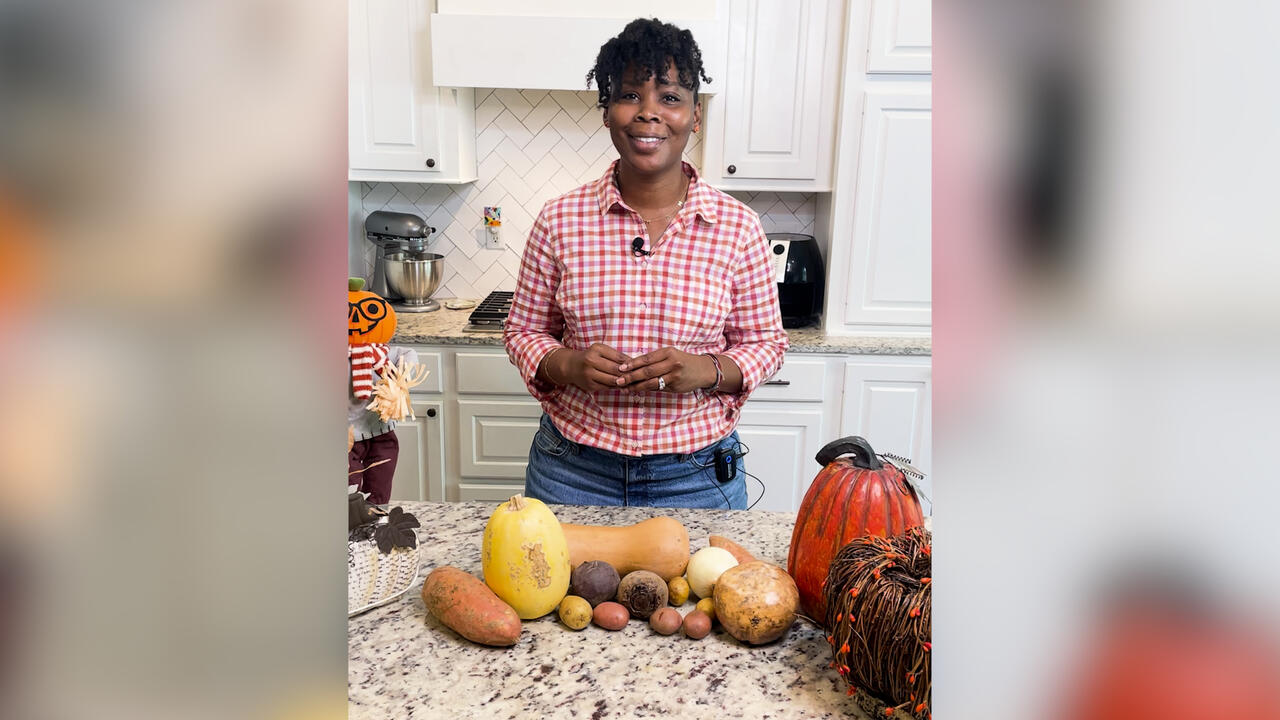 A woman stands in a kitchen with various root vegetables on the counter.