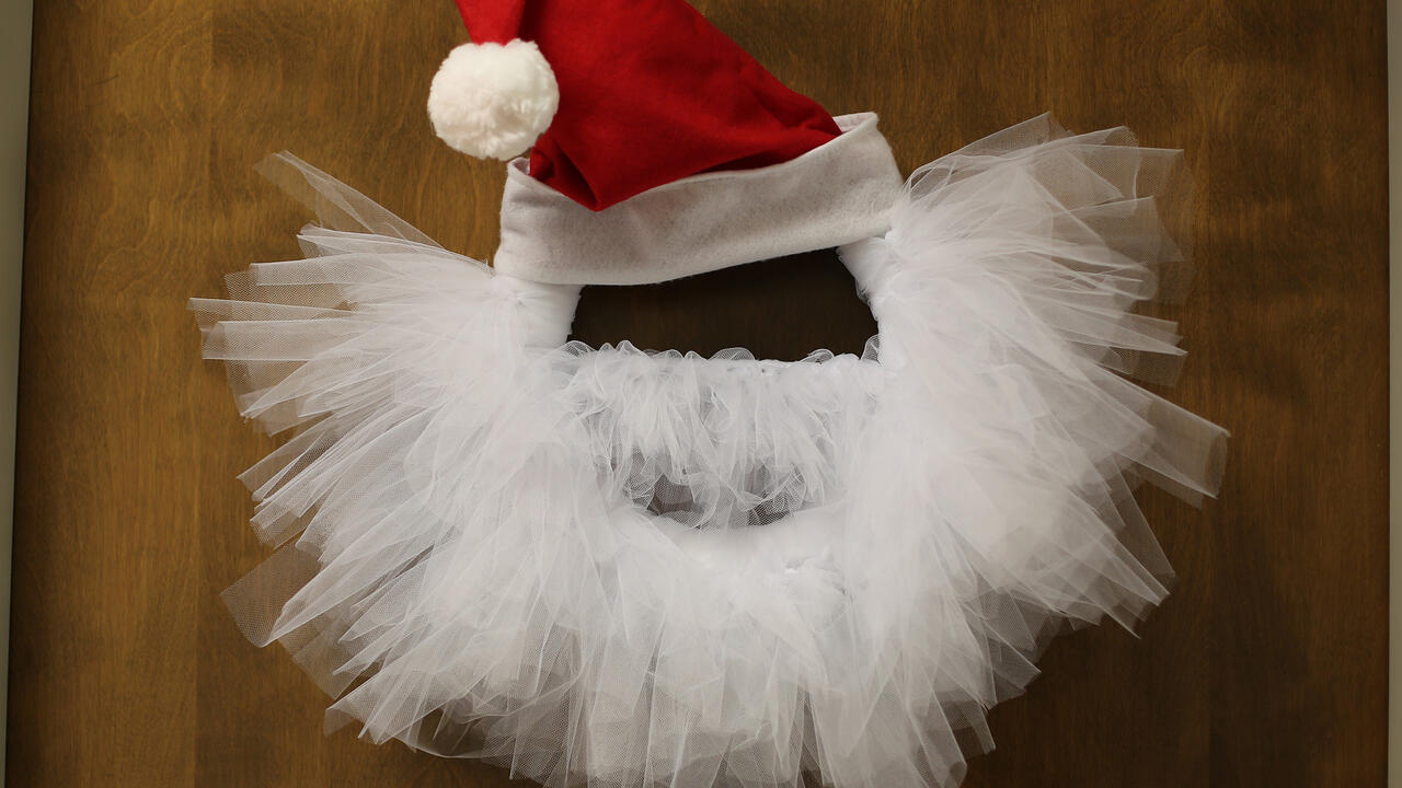 Wreath with Santa hat and white tulle shaped to look like a beard hanging on a wooden door.