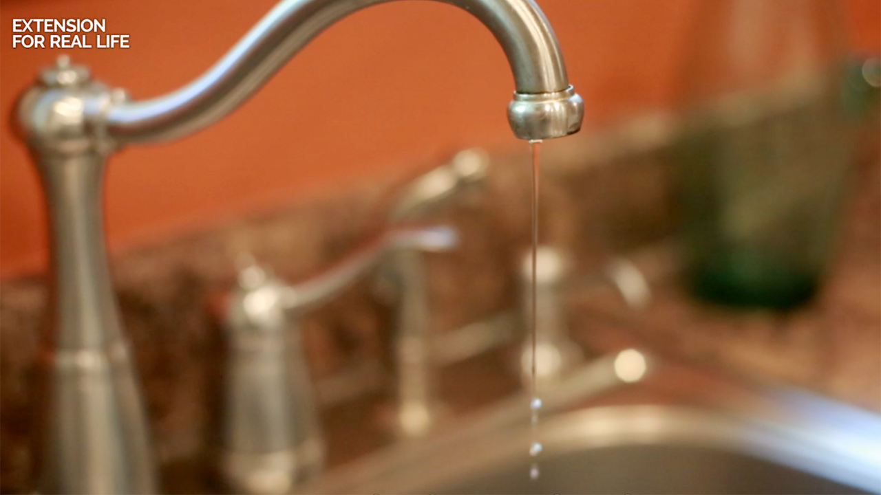 A thin stream of water drips from a stainless steel kitchen sink faucet.