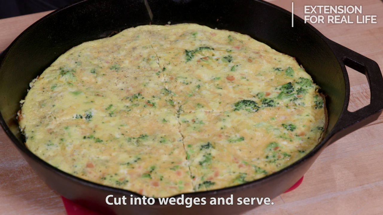A mixture of small broccoli florets, diced carrots, eggs and cheese baked as a frittata in a large black cast-iron skillet.