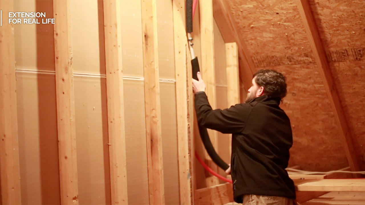 A man fits insulation around water pipes in an unfinished attic of a home.