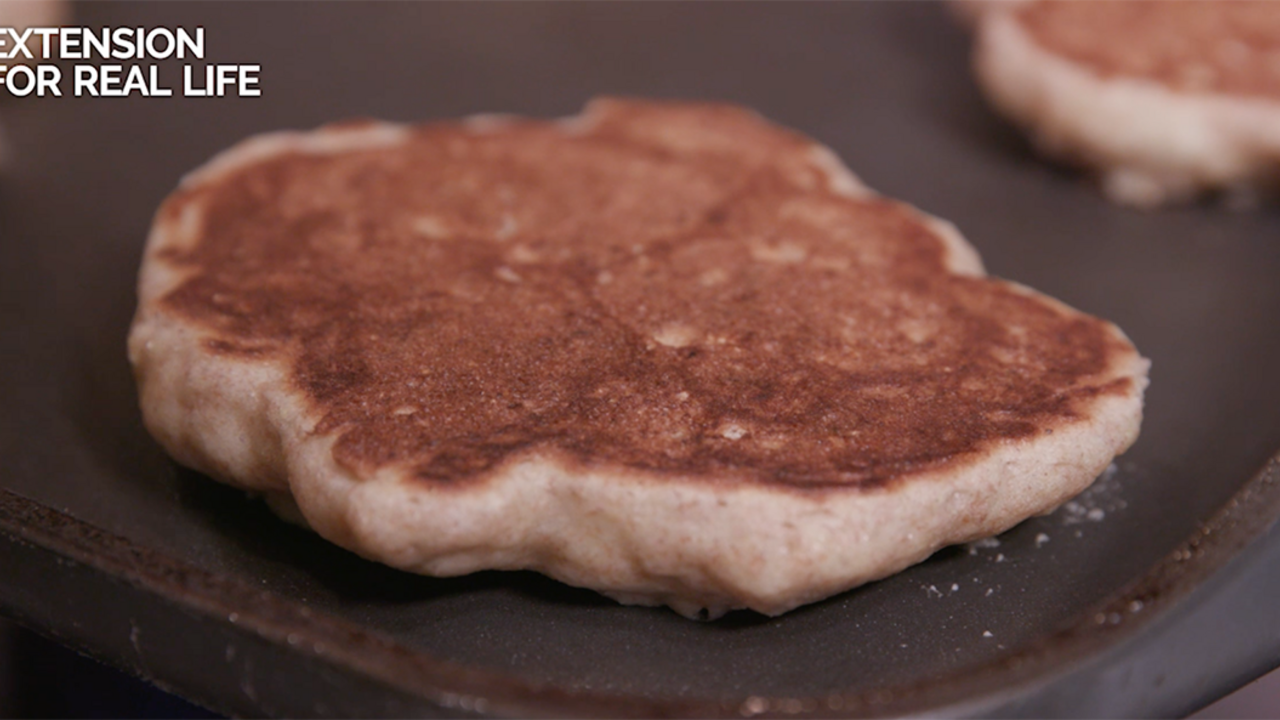 A close-up of a golden brown whole-grain pancake on a nonstick griddle, with parts of two other pancakes blurred in the background. 
